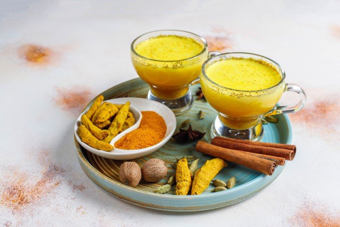 Two cups of golden turmeric milk with turmeric root, cinnamon sticks, cardamom, nutmeg, and turmeric powder arranged on a decorative plate