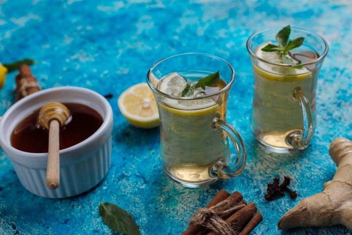 Fresh ginger, lemon, cinnamon sticks, cloves, honey, and two glasses of a warm Jasmine-infusion herbal drink garnished with mint on a blue textured surface.
