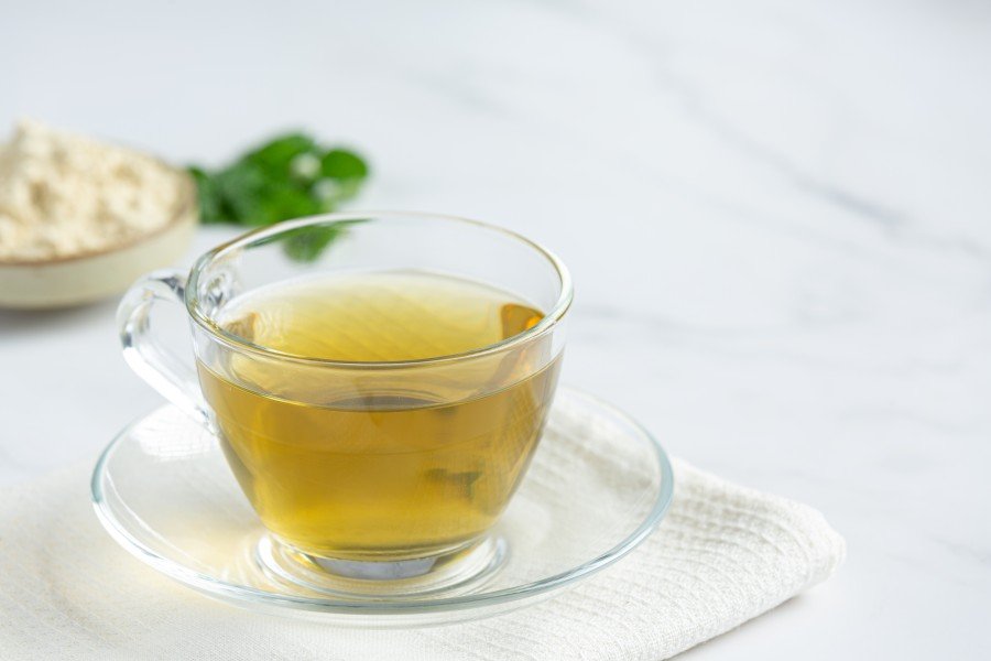 Glass cup of chamomile infusion tea served on a saucer with fresh herbs nearby.