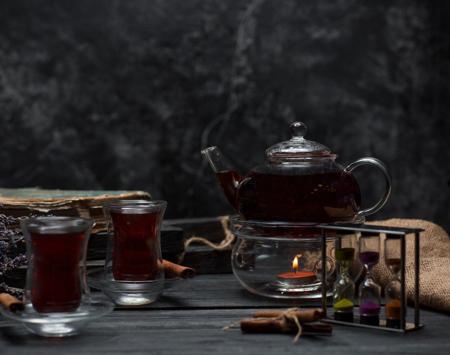 Night Bliss infusion tea served in glass cups and a glass teapot, surrounded by cinnamon sticks, lavender, and an hourglass on a dark rustic table.