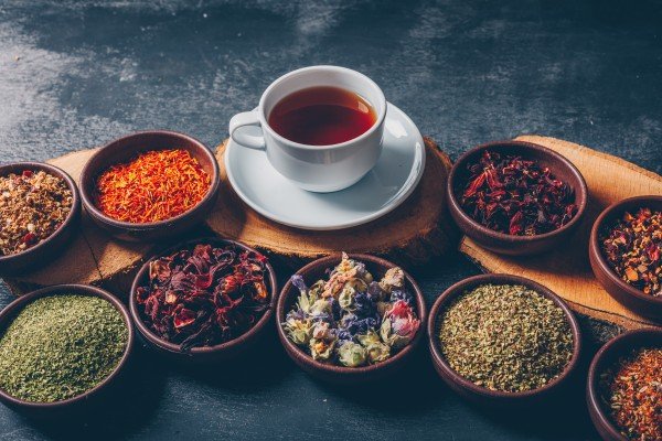 Herbal Tea in a white teacup surrounded by colorful dried herbs, flowers, and loose tea leaves in rustic bowls on a wooden board against a dark textured background.