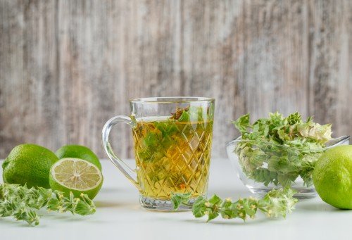Herbal Tea in a glass cup surrounded by fresh green herbs and limes on a white surface, with a rustic wooden background.