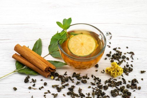 Herbal Tea in a glass cup with fresh lemon, mint leaves, cinnamon sticks, and loose tea leaves on a white wooden table.