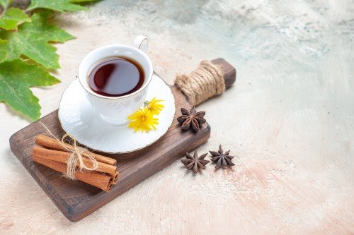 A white teacup filled with dark tea on a saucer, decorated with yellow flowers, cinnamon sticks, and star anise on a wooden tray over a light textured surface.