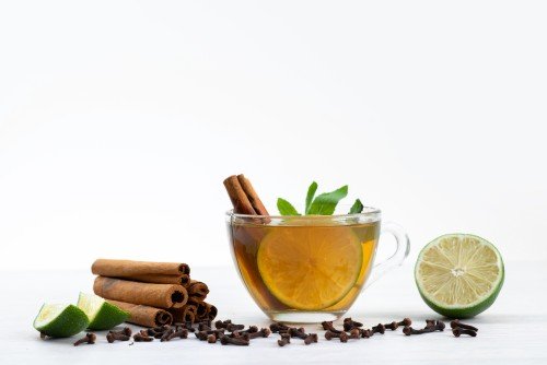 Herbal Tea in a clear glass cup with lemon slices, fresh mint leaves, and cinnamon sticks, surrounded by lime wedges and cloves on a white background.